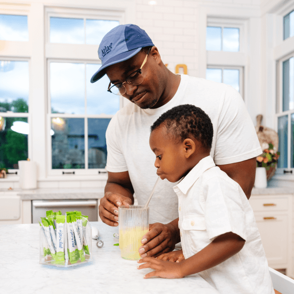 Father helping son in the kitchen. Son is drinking veggie Sips through a straw and glass